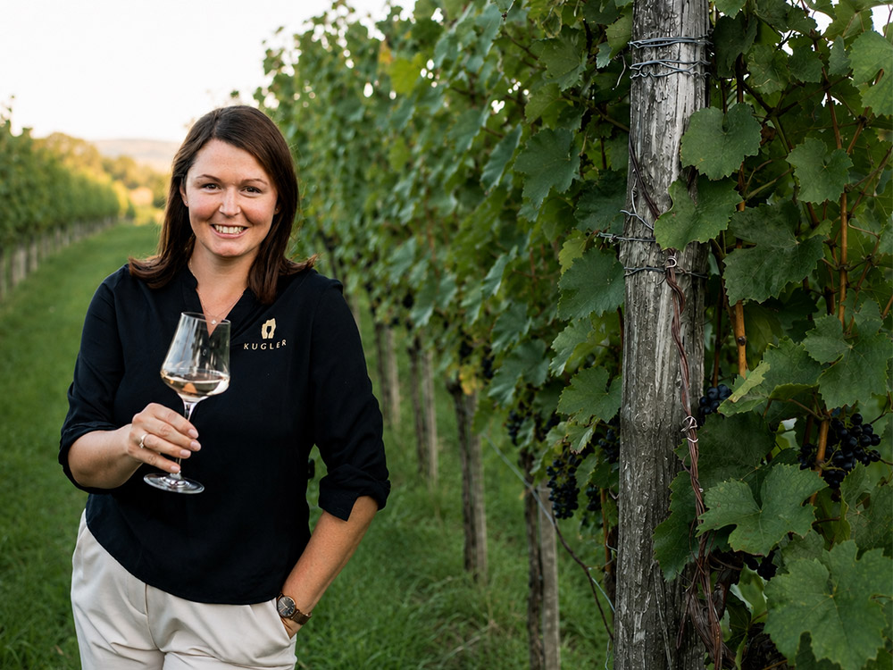 Eine Frau steht in einem Weinberg, hält ein Glas Weißwein in der Hand und lächelt in die Kamera, während zu beiden Seiten Weinreben zu sehen sind.